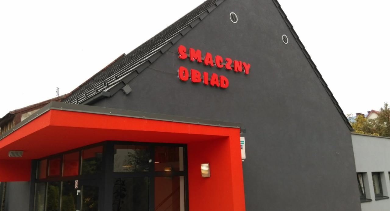 Zewnętrzny widok budynku z napisem 'SMACZNY OBIAD' w czerwonych literach na szarej ścianie, czerwony daszek nad wejściem z przeszklonymi drzwiami.