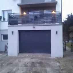 Nowoczesny dom z szarą bramą garażową i balkonem z poziomymi, metalowymi balustradami, oświetlonymi od dołu.