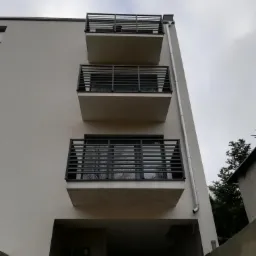 Fasada budynku z trzema balkonami zabezpieczonymi nowoczesnymi, metalowymi balustradami o poziomym układzie szczebli, widok z dołu na tle pochmurnego nieba.