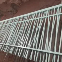 Fragment metalowej konstrukcji z pionowymi prętami o nieregularnym rozmieszczeniu, pomalowanej na jasny kolor, w warsztacie.