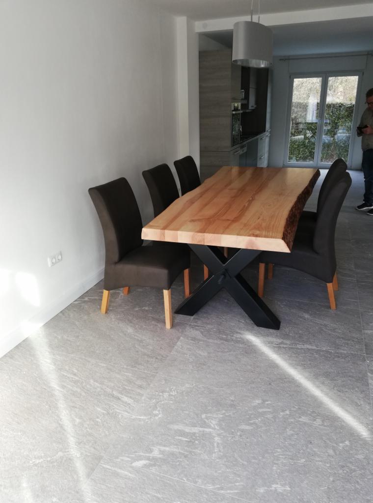 Stół jadalny z blatem z litego drewna o nieregularnych krawędziach, osadzony na czarnej, metalowej podstawie w kształcie litery X, otoczony krzesłami z ciemnoszarą tapicerką w nowoczesnym wnętrzu.