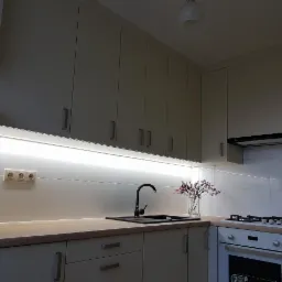 Jasna kuchnia z kremowymi szafkami, czarnym zlewem i oświetleniem LED pod szafkami wiszącymi, uzupełniona o kwiaty w wazonie.