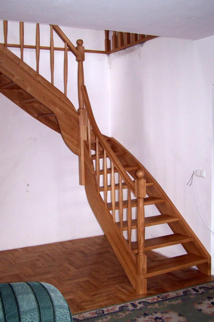 Drewniane schody z zakrętem, balustrada z toczonymi elementami, widoczne parkiet i fragment sofy.