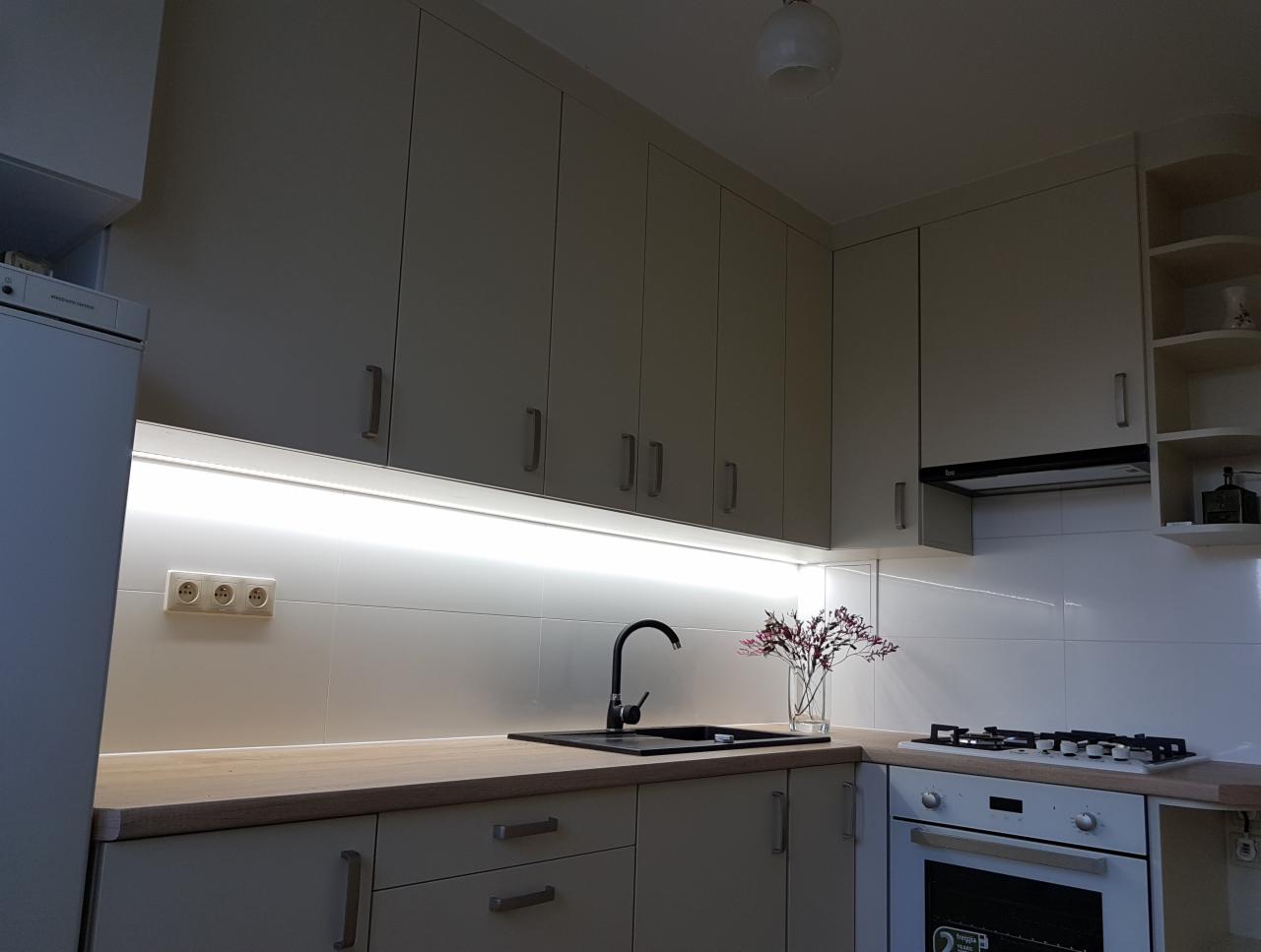 Jasna kuchnia z kremowymi szafkami, czarnym zlewem i oświetleniem LED pod szafkami wiszącymi, uzupełniona o kwiaty w wazonie.