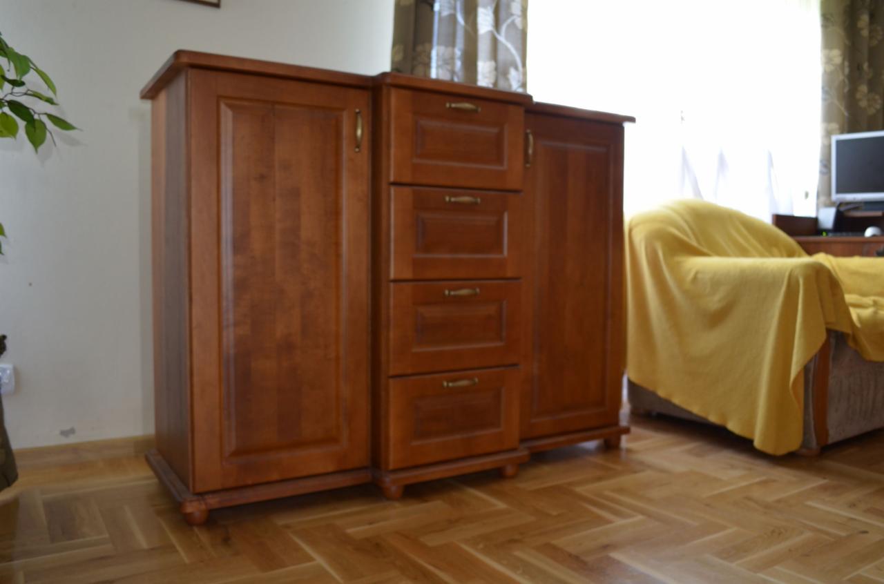 Drewniany mebel z szafkami i szufladami w ciepłym odcieniu brązu, stojący na parkiecie w jodełkę, z widocznym fragmentem rośliny doniczkowej i zasłoniętym oknem w tle.