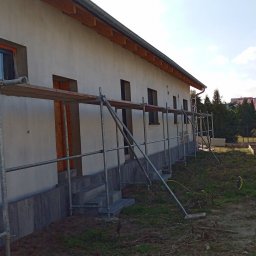 Damian Kopka - Budynek jednorodzinny w trakcie prac elewacyjnych. Widoczne rusztowania, świeżo otynkowana ściana i elementy wykończeniowe okien. W tle zieleń i zabudowa.