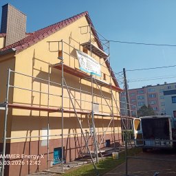 Damian Kopka - Żółta elewacja domu w trakcie malowania, z rusztowaniem i banerem firmy. Widoczny fragment samochodu dostawczego i otoczenie budynku w słoneczny dzień.