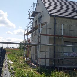 Damian Kopka - Dom jednorodzinny w trakcie prac elewacyjnych, otoczony rusztowaniem. Widoczne świeże tynki i dach z ciemnej dachówki. W tle błękitne niebo z chmurami.