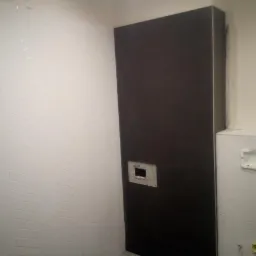 Ściana łazienki wyłożona białymi, wąskimi płytkami w jodełkę, sąsiadująca z pionowym, ciemnym panelem z otworem na instalację, oraz fragmentem ściany z zamontowanym stelażem podtynkowym.