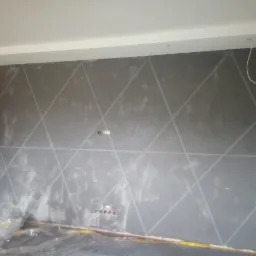 Ściana w szarym odcieniu z geometrycznym wzorem z linii, zabezpieczona folią malarską przy podłodze, widoczne gniazdka elektryczne i wystający kabel.