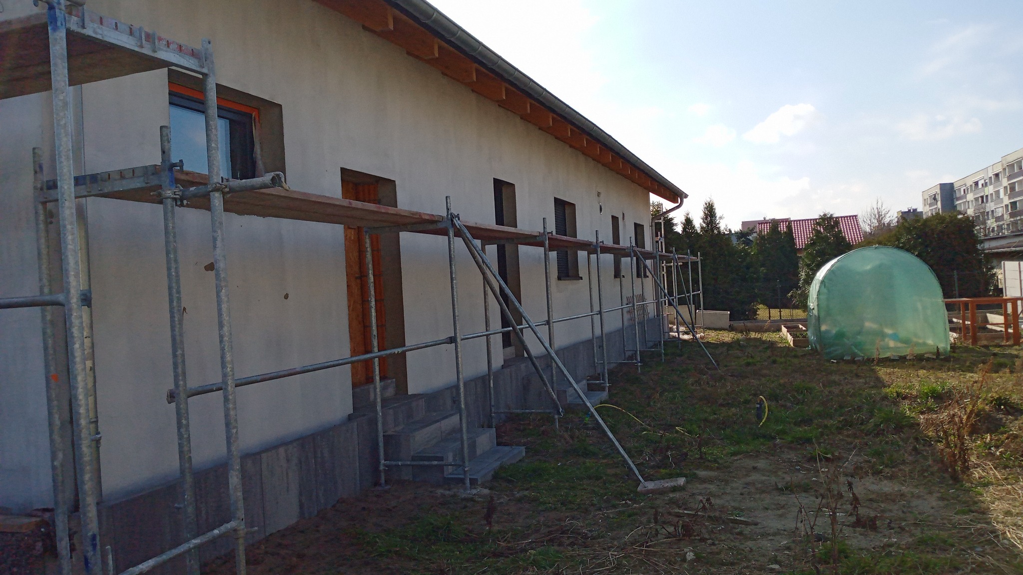 Budynek jednorodzinny w trakcie prac elewacyjnych. Widoczne rusztowania, świeżo otynkowana ściana i elementy wykończeniowe okien. W tle zieleń i zabudowa.