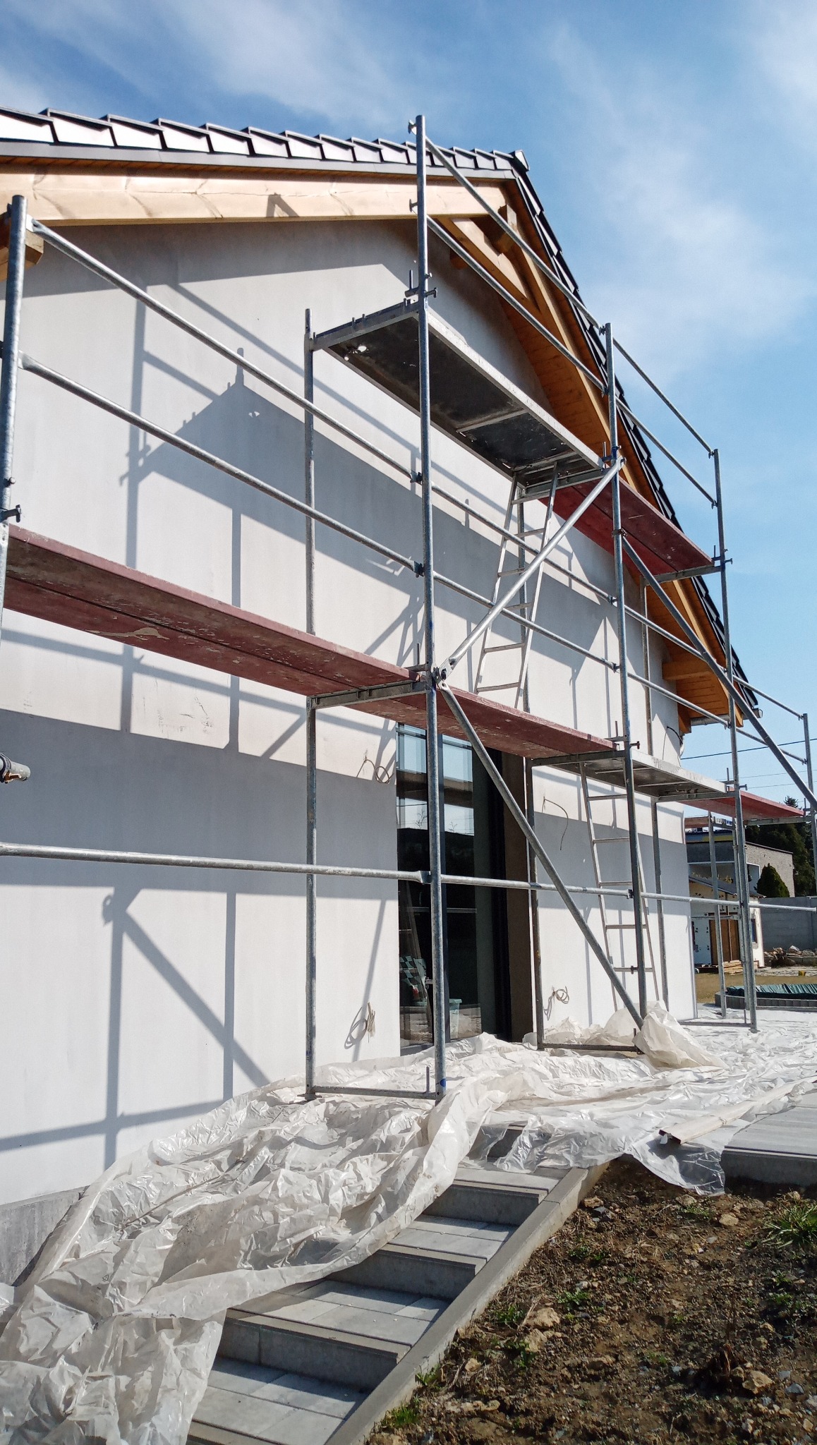 Dom jednorodzinny w trakcie prac elewacyjnych. Widoczne rusztowania, zabezpieczenia folią i fragment otynkowanej ściany w jasnym kolorze. Błękitne niebo w tle.