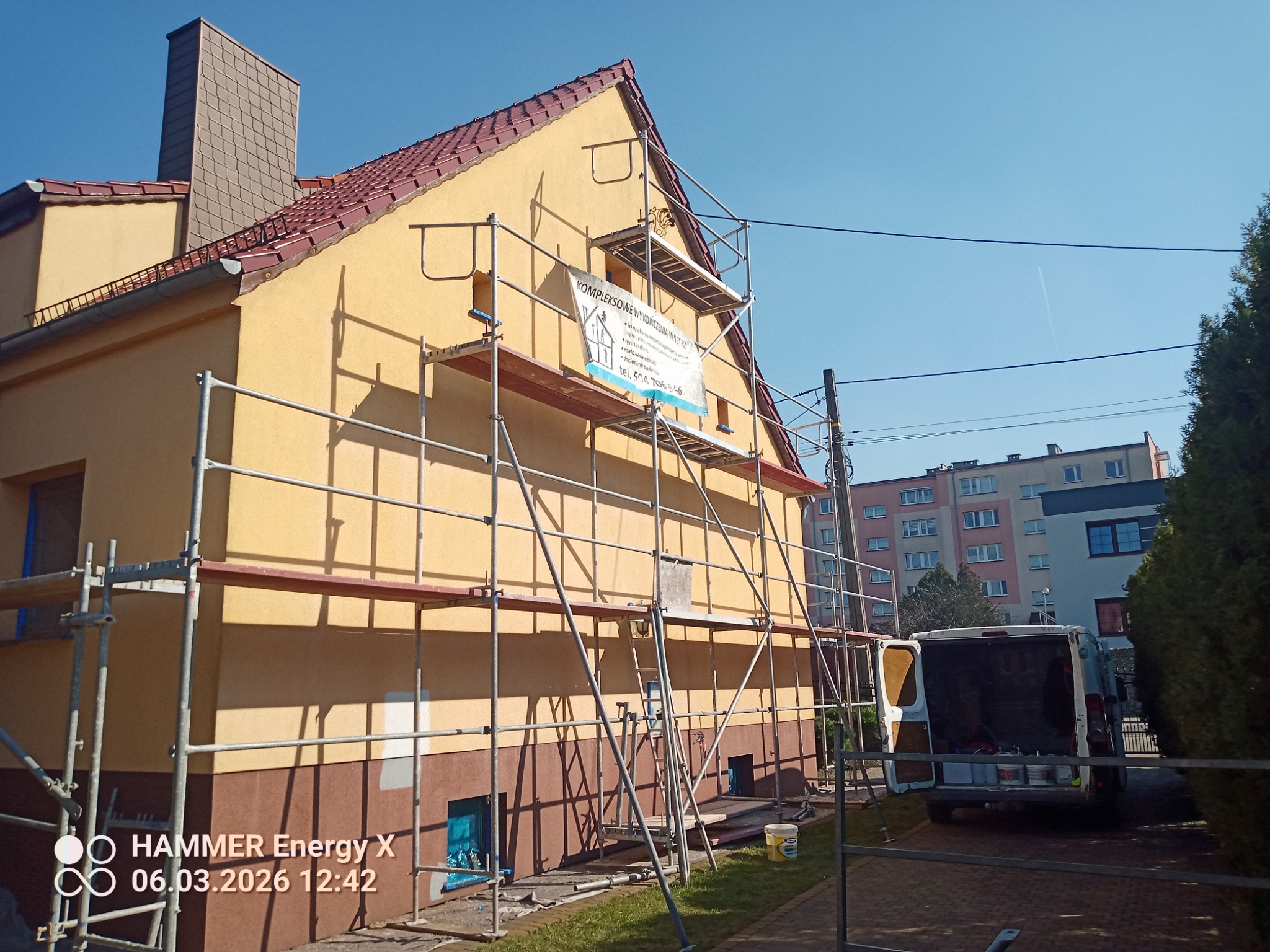 Żółta elewacja domu w trakcie malowania, z rusztowaniem i banerem firmy. Widoczny fragment samochodu dostawczego i otoczenie budynku w słoneczny dzień.