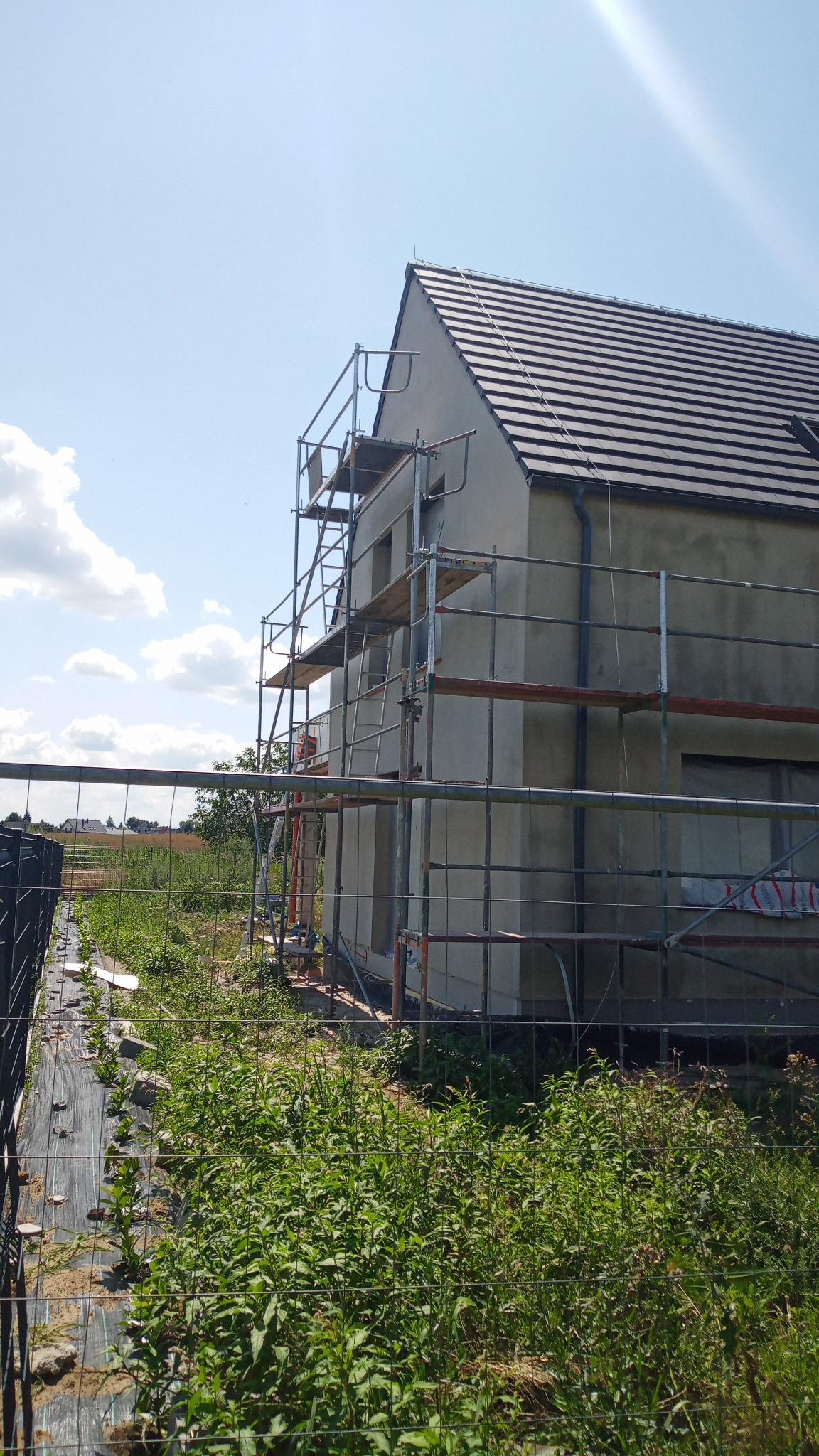 Dom jednorodzinny w trakcie prac elewacyjnych, otoczony rusztowaniem. Widoczne świeże tynki i dach z ciemnej dachówki. W tle błękitne niebo z chmurami.