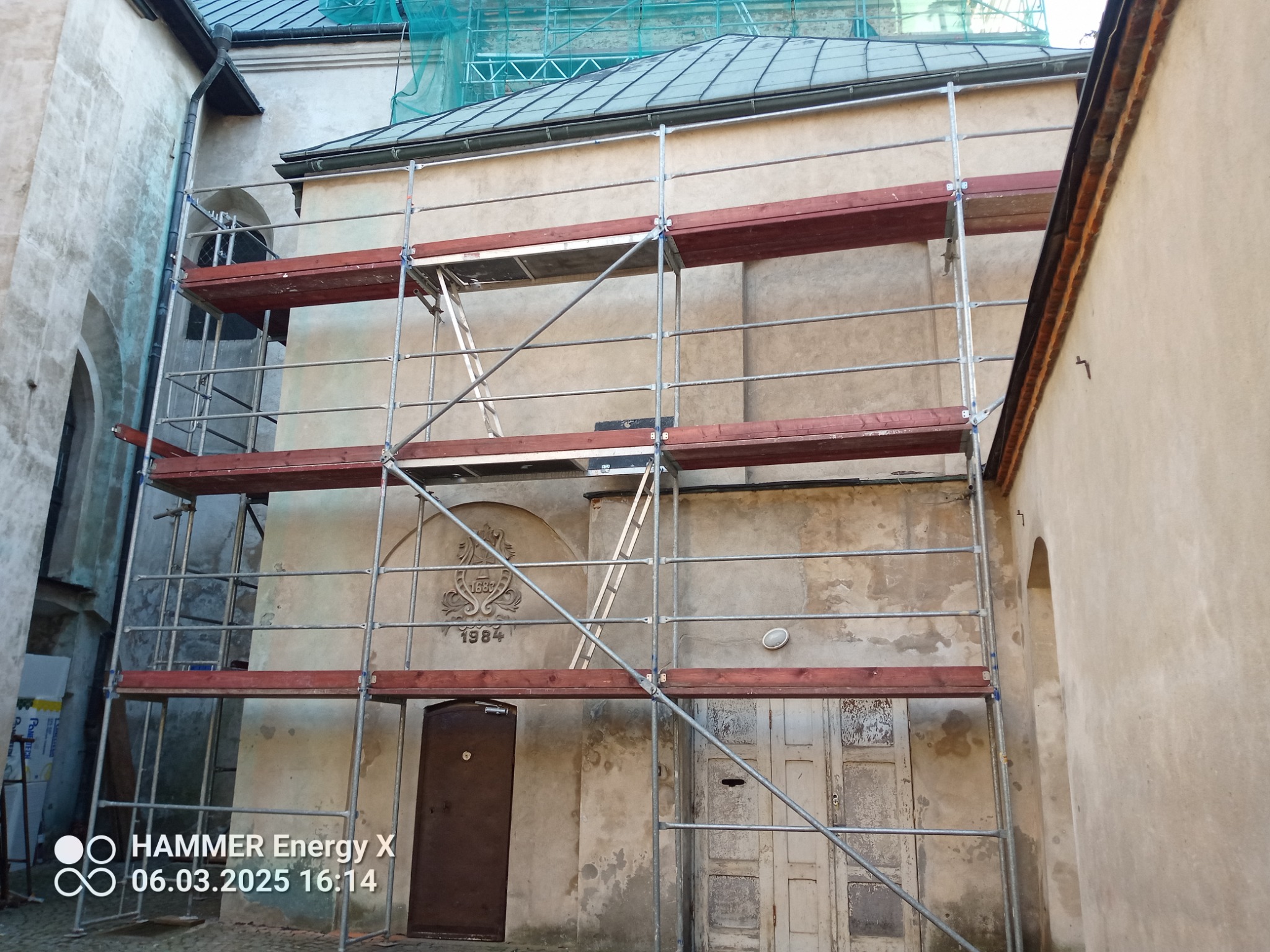 Elewacja budynku w trakcie renowacji, z widocznymi rusztowaniami i dekoracyjnym elementem z datą 1684/1984. Prace konserwatorskie w toku, widoczne ślady napraw.