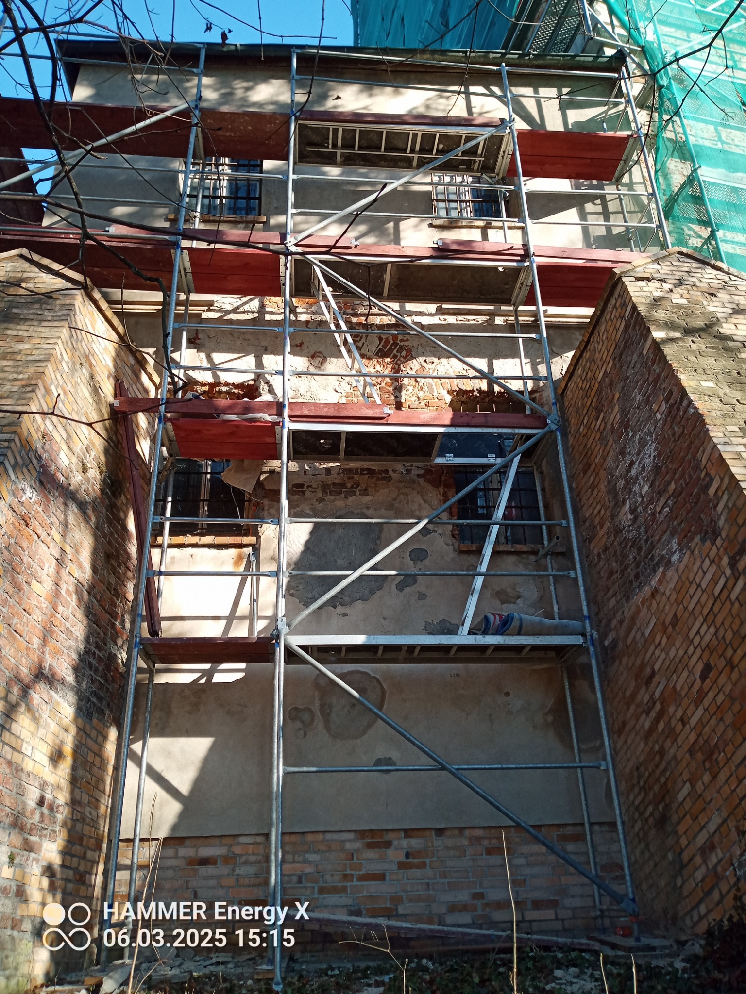 Budynek w trakcie renowacji z rusztowaniem. Widoczne fragmenty starej elewacji i ceglanej ściany. Częściowo zasłonięty siatką ochronną. Ujęcie z dołu.
