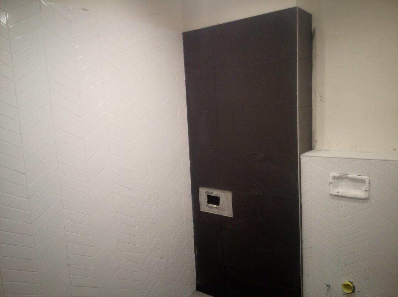 Ściana łazienki wyłożona białymi, wąskimi płytkami w jodełkę, sąsiadująca z pionowym, ciemnym panelem z otworem na instalację, oraz fragmentem ściany z zamontowanym stelażem podtynkowym.