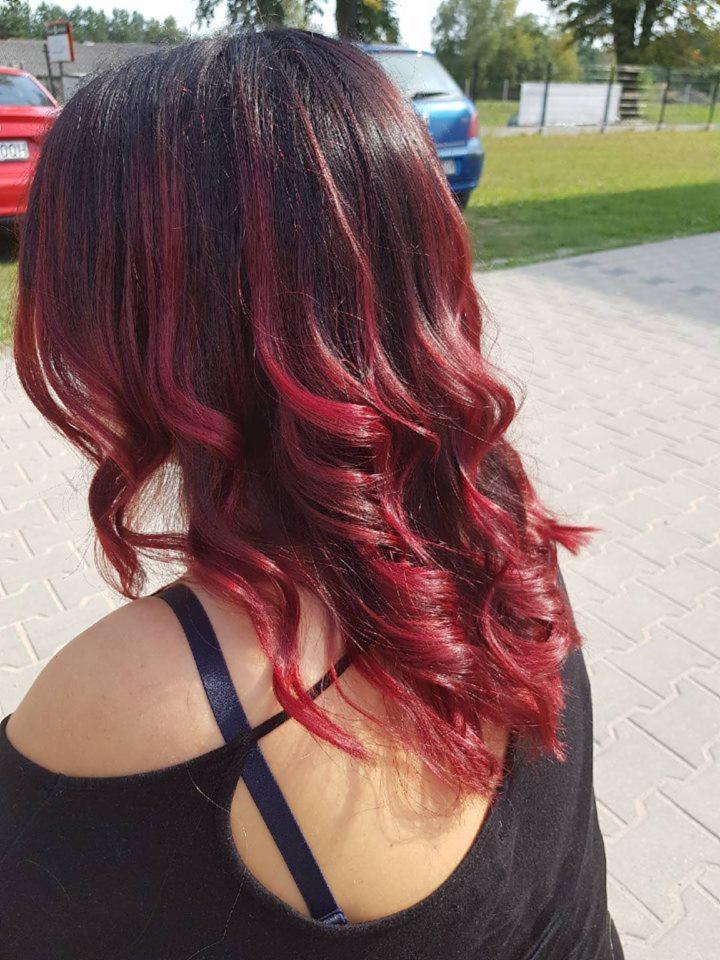 Widok z tyłu na kobiece włosy z ciemnego brązu przechodzące w intensywną czerwień, układające się w luźne fale, na tle chodnika i zaparkowanych samochodów.