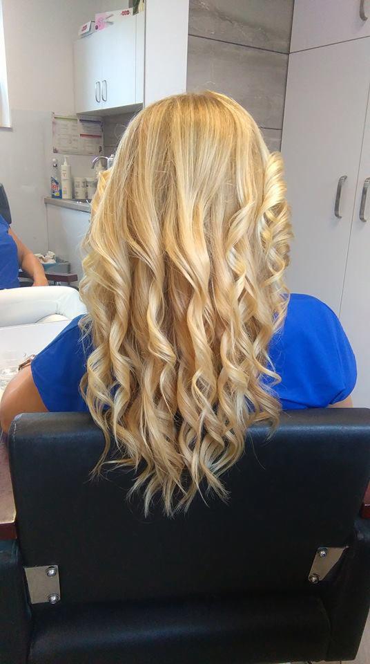 Tył głowy blondynki z długimi, gęstymi lokami w salonie kosmetycznym, widoczny fragment niebieskiej koszulki i czarny fotel.