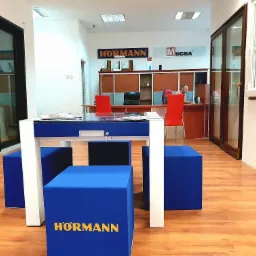 Wnętrze salonu sprzedaży z ekspozycją stolarki otworowej: przesuwne konstrukcje w ciemnych ramach oraz białe okno, widoczne logo firm Hormann i Mucha, w tle pracownik przy biurku.