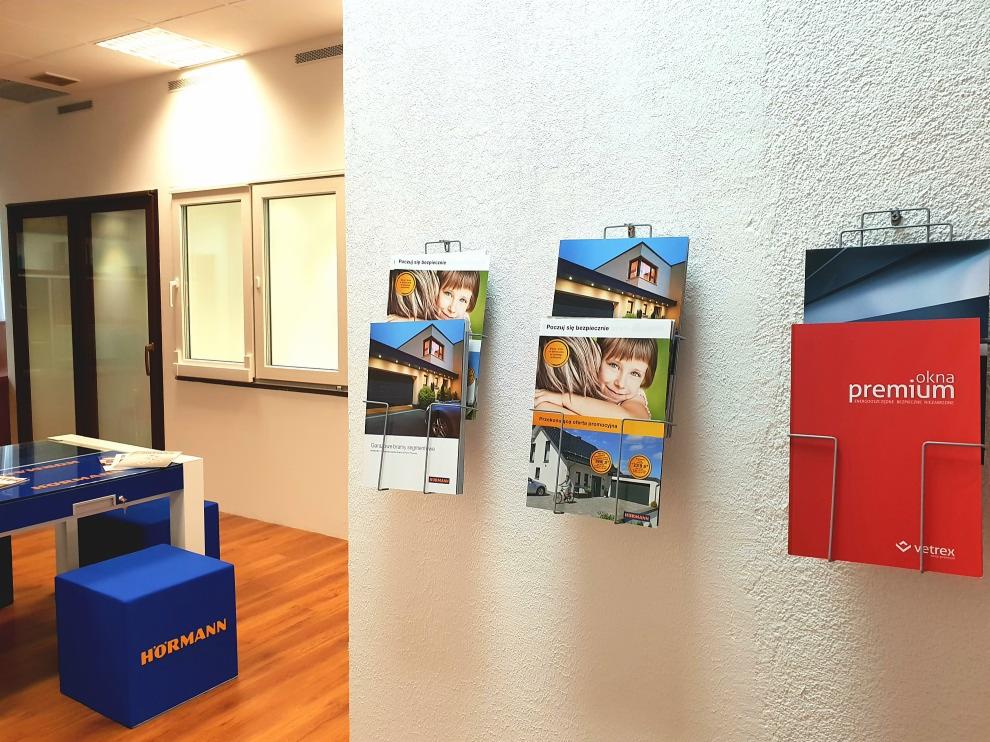 Wnętrze biura z ekspozycją folderów reklamowych producenta stolarki okiennej i drzwiowej, widoczne okna i drzwi w tle.