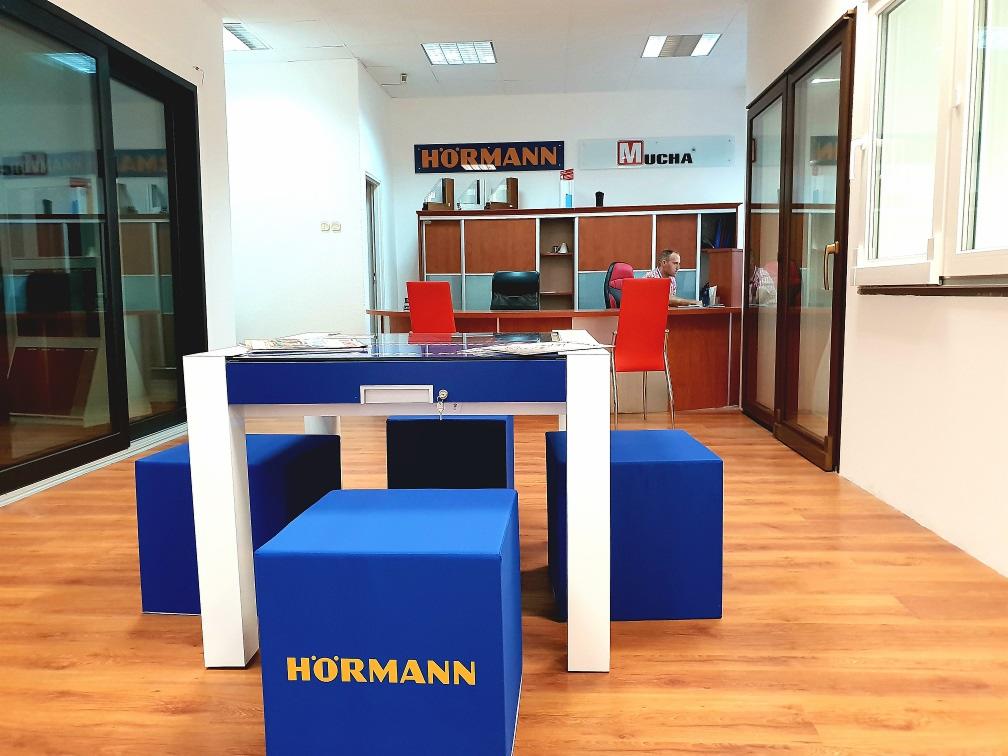 Wnętrze salonu sprzedaży z ekspozycją stolarki otworowej: przesuwne konstrukcje w ciemnych ramach oraz białe okno, widoczne logo firm Hormann i Mucha, w tle pracownik przy biurku.