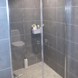 Szara łazienka z kabiną prysznicową, widoczne płytki ceramiczne, półka z kosmetykami i butelka szamponu na podłodze, uchwyt do kabiny.