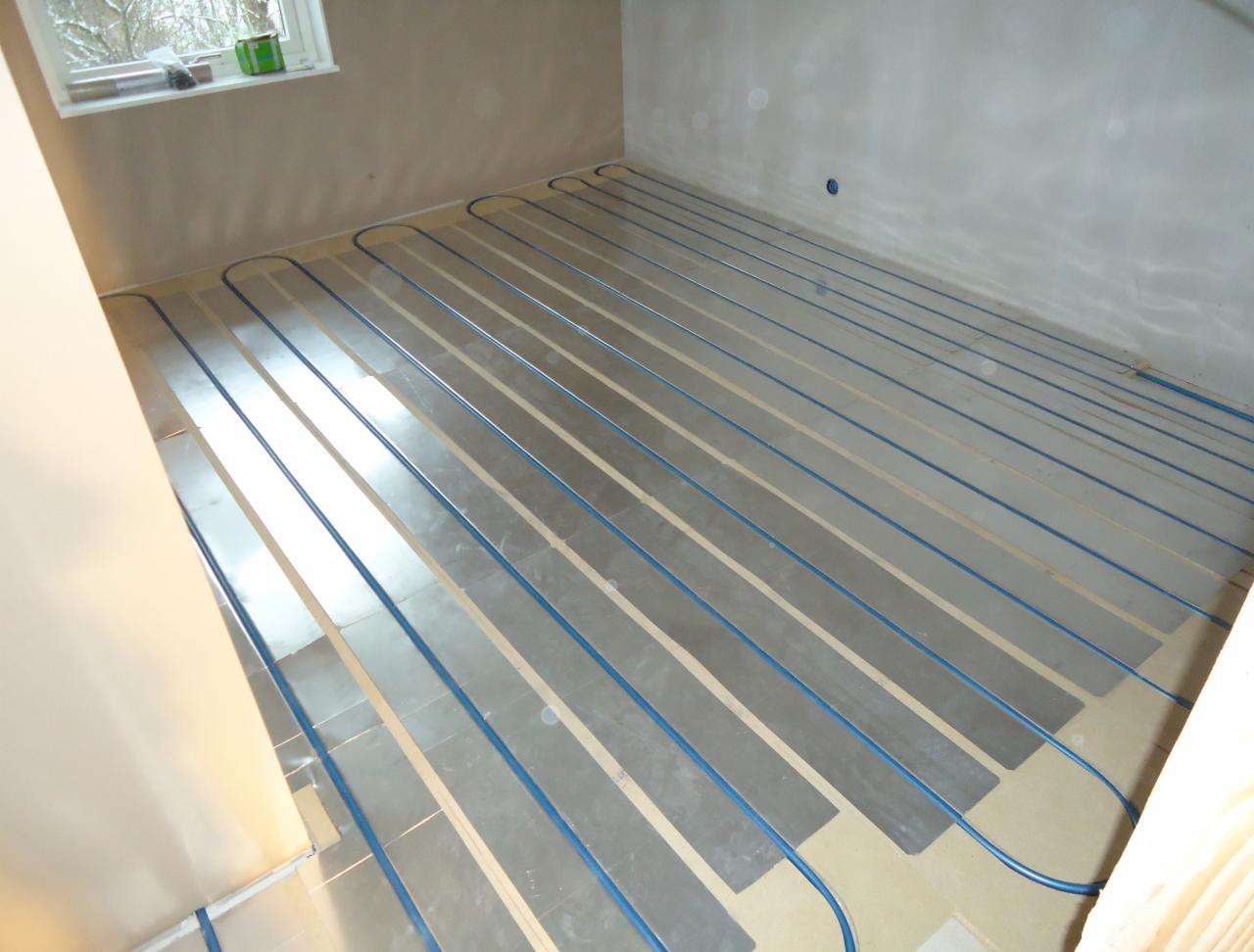 Instalacja ogrzewania podłogowego z niebieskimi rurkami na srebrnych panelach izolacyjnych w surowym wnętrzu pokoju z oknem.
