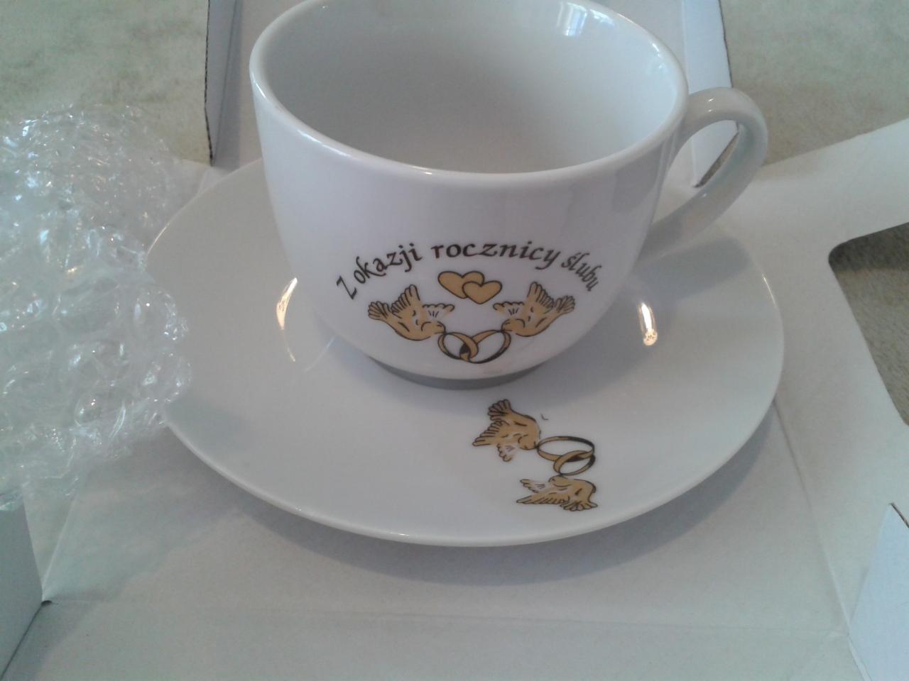 Biała filiżanka i spodek z nadrukiem gołębi, obrączek i napisem 'Z okazji rocznicy ślubu', zapakowane w pudełko z folią bąbelkową.