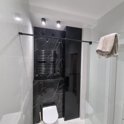 Nowoczesna łazienka z czarno-białymi płytkami imitującymi marmur, podwieszana toaleta, grzejnik drabinkowy i kabina prysznicowa ze szklanymi drzwiami. Minimalistyczny design.