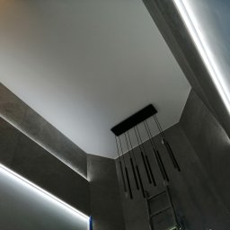 Krasa Krystian - Nowoczesny sufit podwieszany z oświetleniem LED i minimalistyczną lampą wiszącą. Szare płytki na ścianach. Widoczny fragment drabiny.