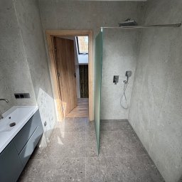 Krasa Krystian - Nowoczesna łazienka na poddaszu z szarymi płytkami imitującymi lastryko na ścianach i podłodze. Widoczna kabina walk-in z deszczownicą, minimalistyczna szafka umywalkowa i drewniane drzwi.