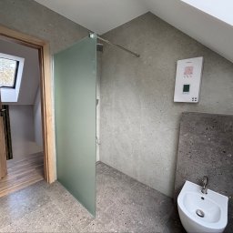 Krasa Krystian - Nowoczesna łazienka z kabiną prysznicową walk-in z matową szybą, bidetem i ściennym podgrzewaczem wody. Szare, nakrapiane płytki na ścianach i podłodze. Otwarte drewniane drzwi do pokoju z oknem...