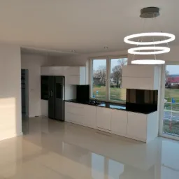 Nowoczesna, minimalistyczna kuchnia z białymi szafkami, czarnym blatem i okrągłą, trzypoziomową lampą LED. Widok na zewnątrz przez duże okna, z widoczną drogą i domami w oddali. Błyszcząca podłoga...