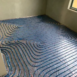 Niebieskie rury ogrzewania podłogowego ułożone spiralnie na srebrnej folii izolacyjnej z czerwoną siatką, widok z góry w surowym wnętrzu z oknem.