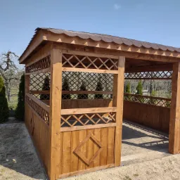 Drewniana altana z kratownicowymi ścianami i dachem z gontu bitumicznego, widok z zewnątrz na tle ogrodu z tujami i drzewami owocowymi.