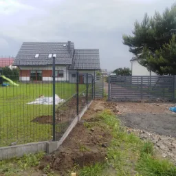 Nowoczesne ogrodzenie panelowe z podmurówką betonową, widok na dom z szarą dachówką i zielony trawnik z placem zabaw w tle.