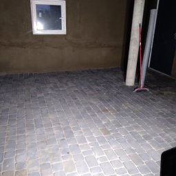 F.U. Benio Benedykt Łącki - Surowa ściana z oknem i ułożona kostka brukowa na podłodze w pomieszczeniu w trakcie remontu, widoczny betonowy słup i rura.