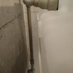 Szare rury hydrauliczne prowadzące do białego zbiornika, zamontowane na surowej, betonowej ścianie z widocznymi śladami po wierceniu i mocowaniach.