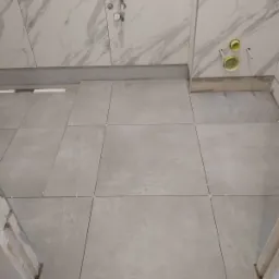 Układanie szarych płytek podłogowych w łazience, widoczne krzyżyki dystansowe, marmurowe płytki ścienne i instalacja hydrauliczna w tle.