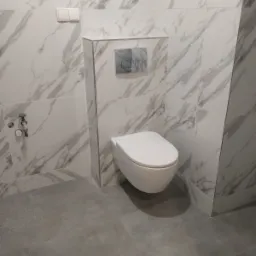 Podwieszana toaleta w wykończonej łazience z marmurowymi płytkami na ścianach i szarą podłogą z płytek, widoczny przycisk spłuczki w kolorze chromu i zawór dopływu wody.