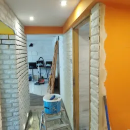 Korytarz w trakcie odświeżania: pomarańczowa ściana kontrastuje z białą imitacją cegły, wiadro z farbą na drabince, widoczne narzędzia malarskie i zabezpieczenia podłogi.