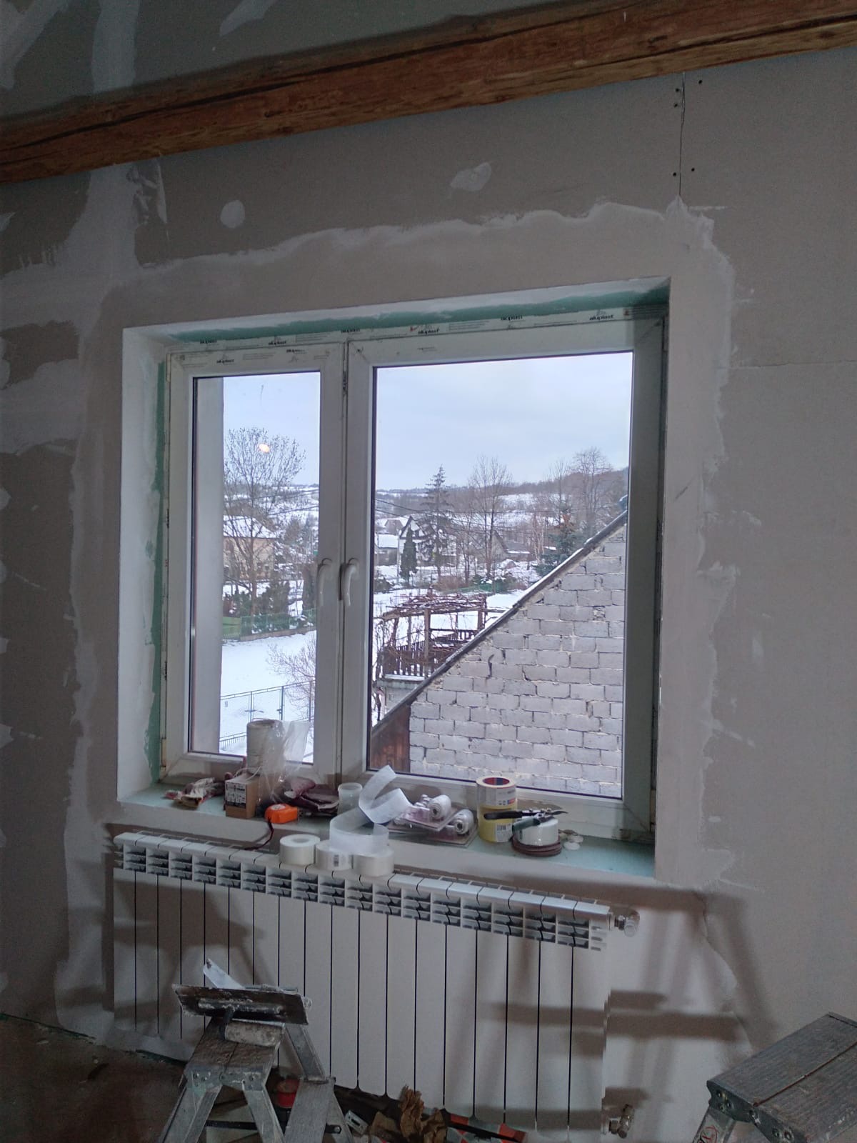Pokój w trakcie szpachlowania ścian wokół okna, widoczne narzędzia na parapecie, grzejnik i drewniane belki pod sufitem.