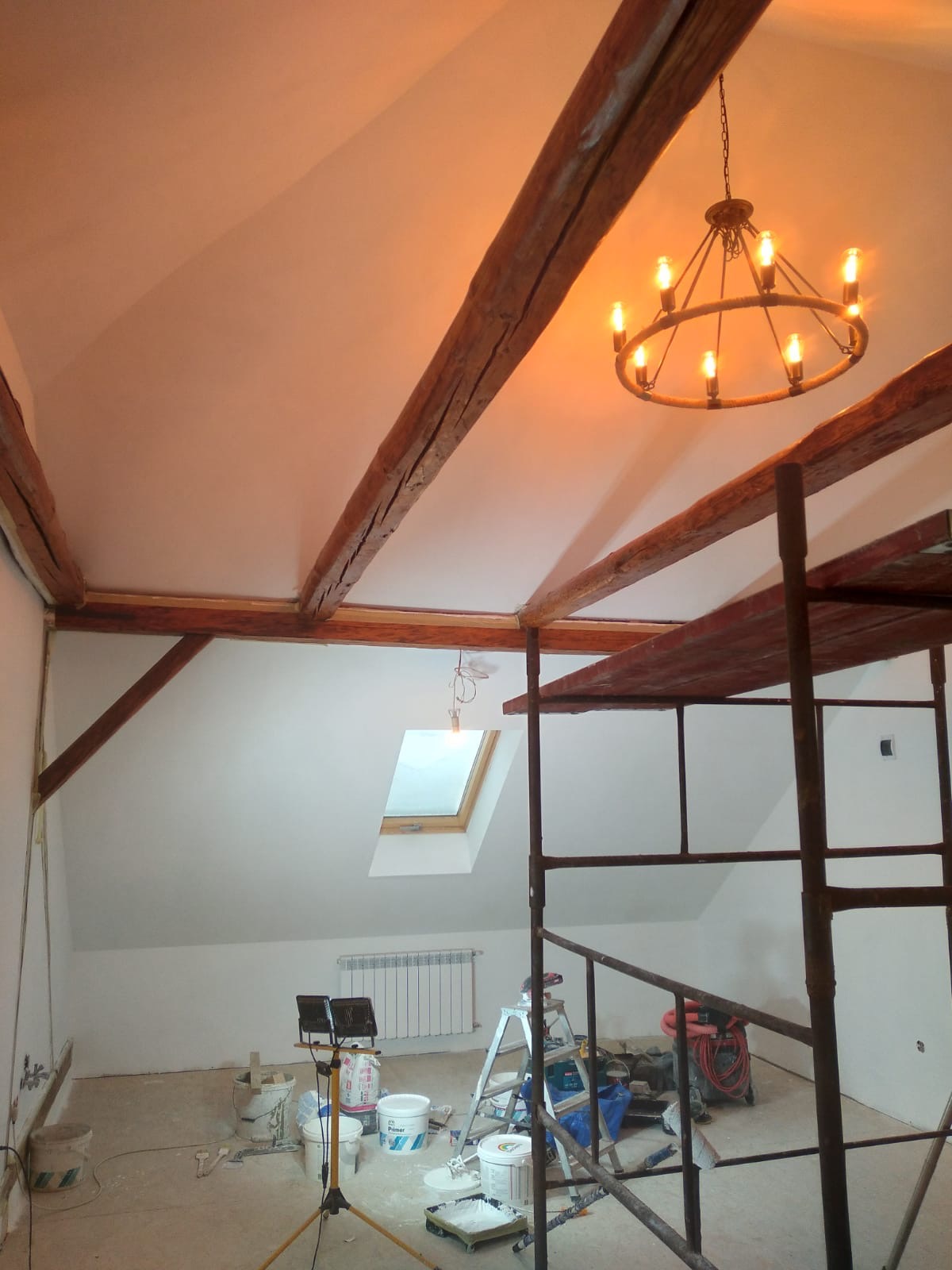 Poddasze w trakcie prac wykończeniowych: widoczne drewniane belki stropowe, białe ściany, okno dachowe, metalowy żyrandol w stylu retro, rusztowanie oraz narzędzia malarskie.