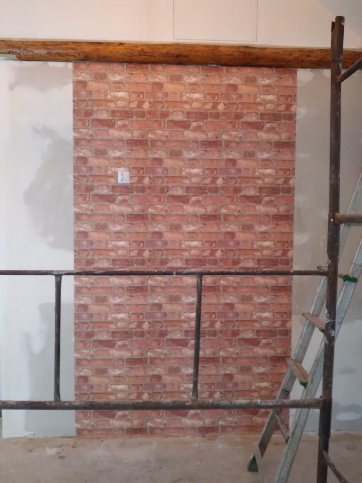 Fragment ściany z imitacją cegły, widoczne rusztowanie i drabina, surowy drewniany belka stropowa, ściana po bokach niezakończona, gniazdko elektryczne na ceglanej ścianie.