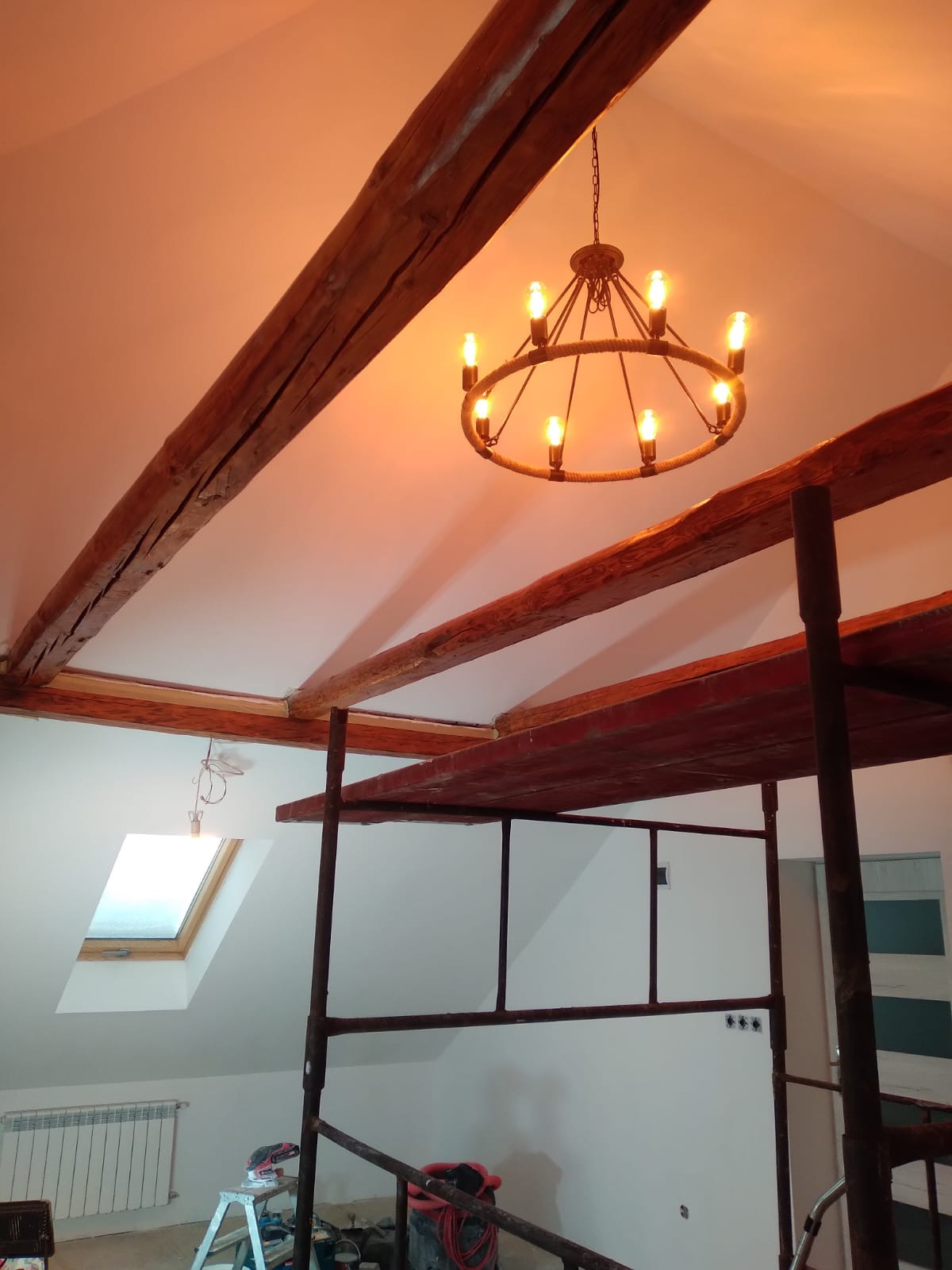 Klatka rusztowania w trakcie prac przy wykończeniu poddasza ze skośnym sufitem, widoczne drewniane belki stropowe, okno dachowe, grzejnik i wiszący żyrandol z żarówkami imitującymi świece.