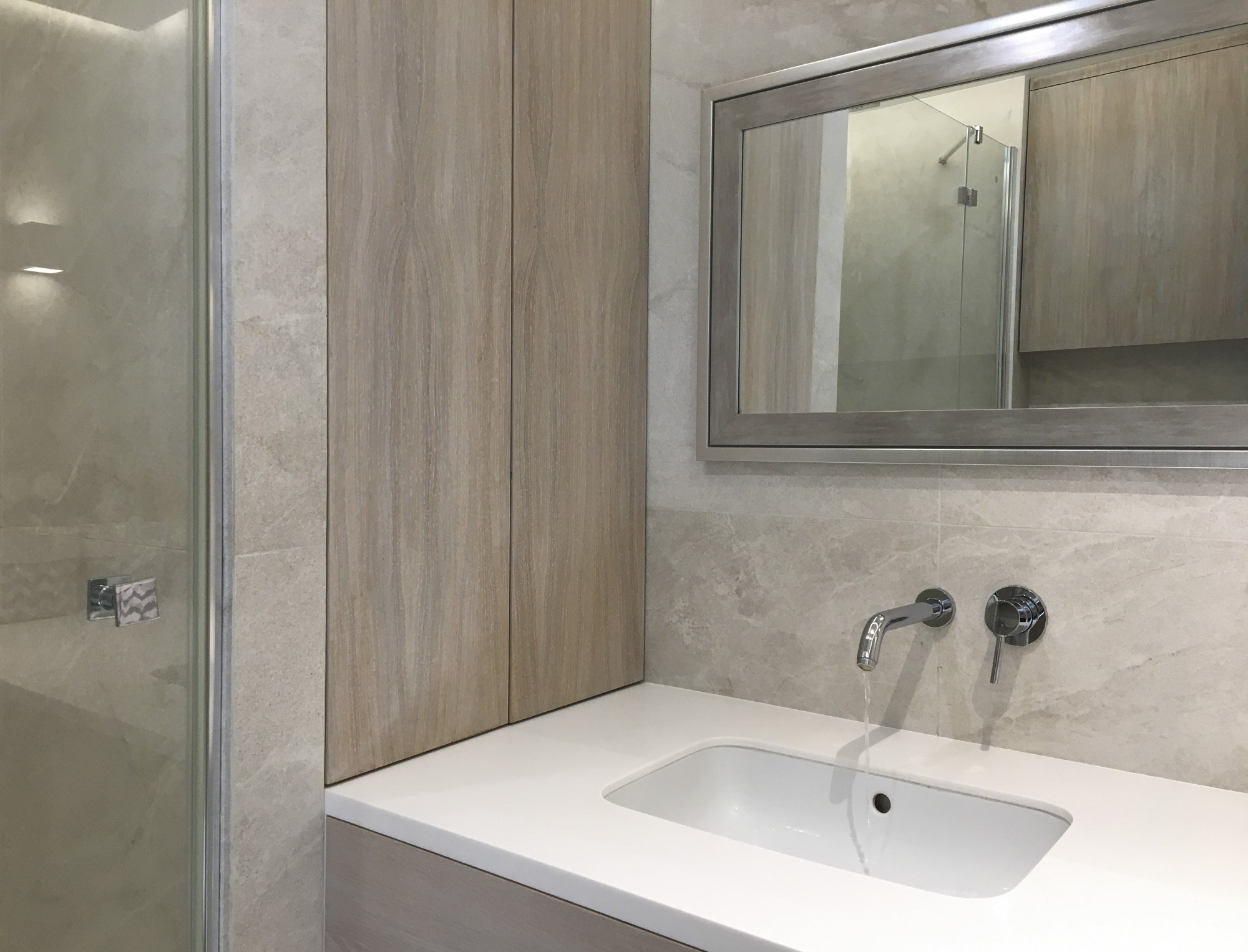 Nowoczesna łazienka z umywalką wpuszczoną w blat, bateria ścienna z włączonym strumieniem wody, lustro w ramie i szafka z frontami imitującymi drewno, całość wykończona płytkami w odcieniach szarości.