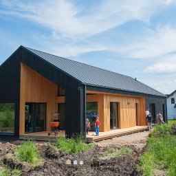 Hygge Hus Sp. z o.o. - Nowoczesny dom szkieletowy z czarną elewacją i drewnianymi panelami, z tarasem i grillem, częściowo zagospodarowany teren wokół domu, widoczni ludzie.