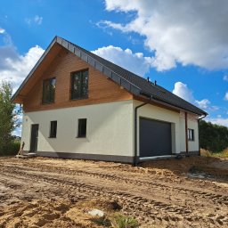 Hygge Hus Sp. z o.o. - Nowoczesny dom z drewnianą elewacją na poddaszu, grafitowa brama garażowa, teren wokół domu w trakcie prac ziemnych, błękitne niebo z białymi chmurami.