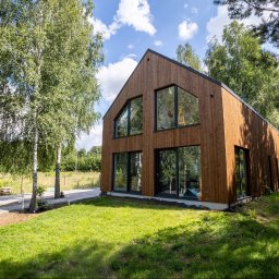 Hygge Hus Sp. z o.o. - Nowoczesny dom z drewnianą elewacją i dużymi oknami, otoczony zielonym trawnikiem i drzewami brzozy w słoneczny dzień.